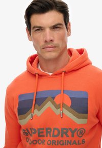 Sudadera naranja con cordón y un diseño gráfico que presenta montañas y un sol en tonos azules y tierras con el texto "SUPERDRY".