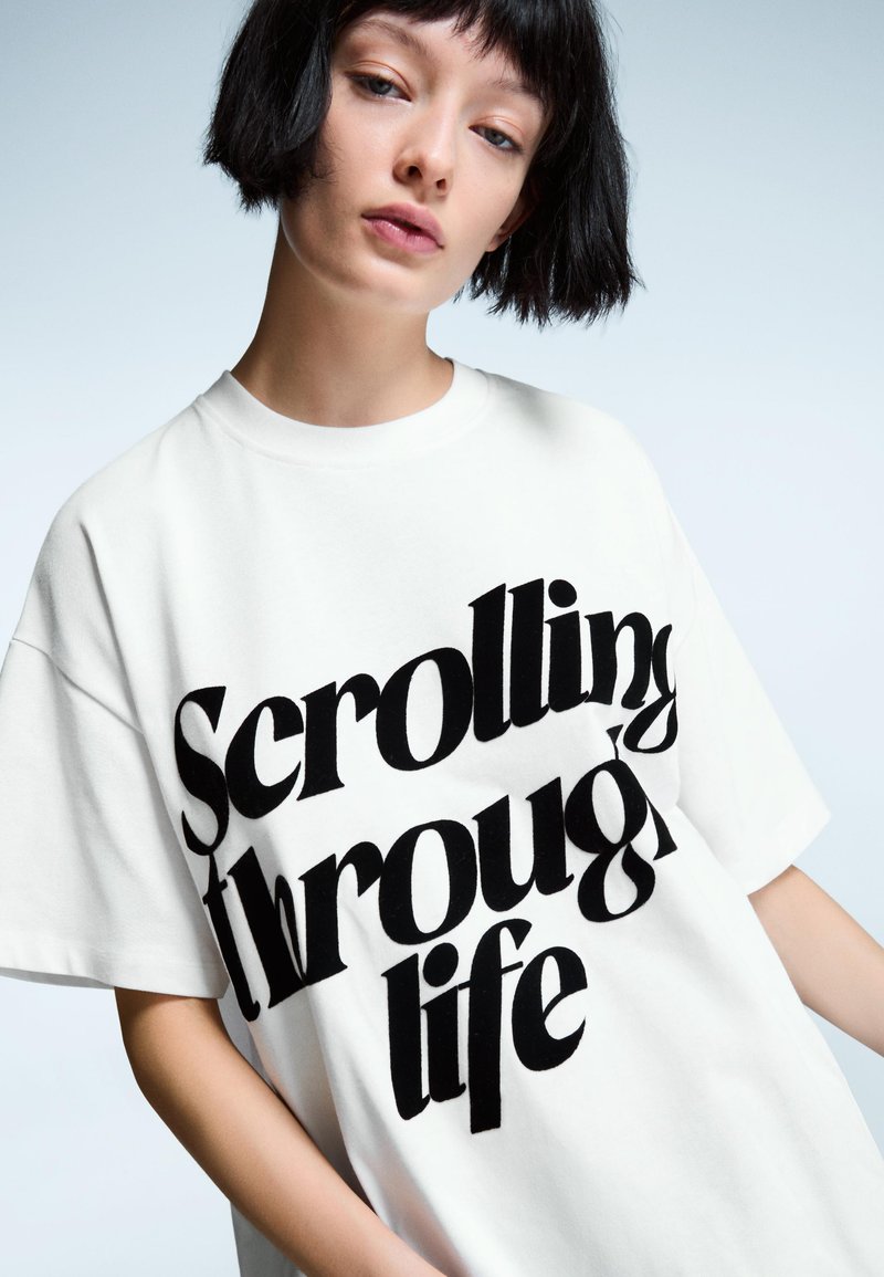 Oversized weißes T-Shirt aus Baumwolle, mit großen schwarzen Buchstaben, die "Durch das Leben scrollen" in einer fetten, stilisierten Schriftart lesen.
