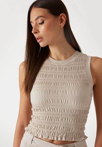 Haut sans manches beige avec des détails de smocks horizontaux, texture côtelée et design ajusté. Encolure ras du cou et ourlet froncé.