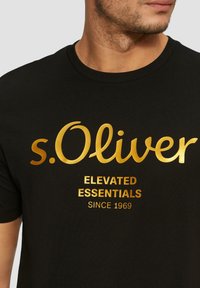 Černé bavlněné tričko s zlatým fóliovým potiskem, na kterém je napsáno "s.Oliver ELEVATED ESSENTIALS SINCE 1969" v klasickém designu.