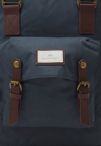 Navy canvas rugzak met een voorzak met een crèmekleurig label, leren accenten in bruin en details in messing.