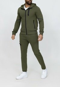 Survêtement de sport vert olive avec un hoodie zippé, des accents noirs et des poches latérales. Associé à des baskets blanches pour un contraste.