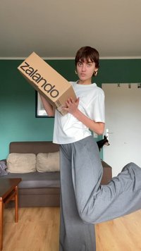 Zalando škatla, ki jo drži oseba v beli majici in sivih širokih hlačah, stoji na eni nogi, v ozadju pa sta kavč in vrata.