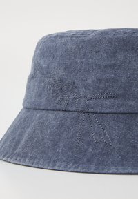 Adjungshatt av denim i mørkeblått med bred skygge. Har subtil blomsterbroderi langs båndet, som viser tekstur og håndverk.