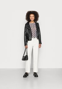 Jaqueta de couro preta, blusa estampada em tons de marrom, calças largas brancas, bolsa preta e sapatos de salto grosso. Fundo neutro.