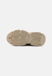 Suela de zapatilla beige con goma texturizada, que presenta patrones de relieve distintos y el nombre de la marca en relieve. Punta redondeada y construcción robusta.