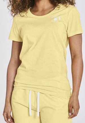 Kobieta ubrana w żółtą koszulkę z krótkim rękawem i pasujące żółte spodnie z białymi sznurkami, stojąca na jednolitym tle.