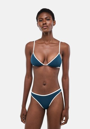 Femme debout, portant un bikini bleu foncé avec des bordures blanches, sur un fond gris clair uni, face à l'objectif avec une expression neutre.