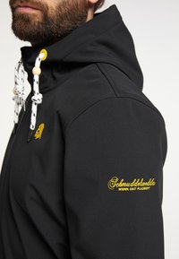 Jachetă neagră din softshell cu glugă, dotată cu șnururi albe și negre cu model și text brodat galben pe mânecă și piept.