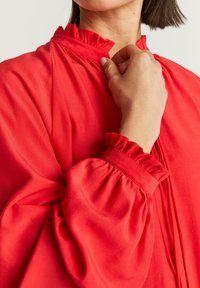 Röd blus med volangkrage och manschetter, med långärmar och en jämn textur. Designen inkluderar vertikala veck.