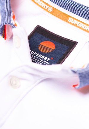 Biała koszulka polo z kołnierzykiem w niebiesko-pomarańczowe paski, czarną metką z logo słońca oraz pomarańczowymi akcentami na listwie guzików.