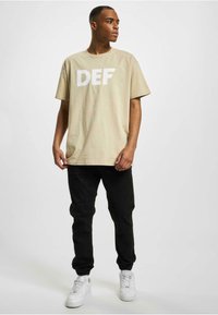 Beige bomull t-skjorte med stort hvitt "DEF" trykk, kombinert med svarte joggebukser og hvite sneakers. Modellen står foran en nøytral bakgrunn.