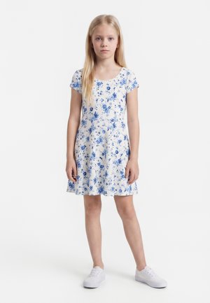 Pige står, iført en hvid kortærmet kjole med blåt blomsterprint og hvide sneakers, vendt mod fronten mod en ensfarvet baggrund.