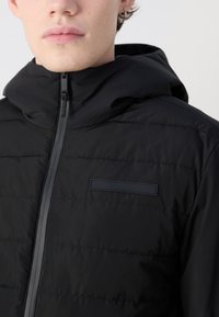 Giacca puffer nera con cappuccio, realizzata in un tessuto liscio e texturizzato, con una zip frontale e un piccolo logo Calvin Klein sul petto.