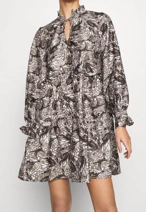 Žena nosí dlouhý rukáv, šaty po kolena s abstraktními vzory v hnědé, černé a bílé barvě a výstřihem na klíč s mašlí.