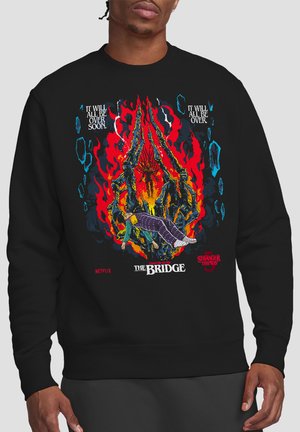 Schwarzer Pullover mit flammenden skelettartigen Händen, die eine schwebende Person in lila Schlafanzug halten. Der Text lautet: "Es wird bald alles vorbei sein", "Die Brücke" und "Stranger Things".