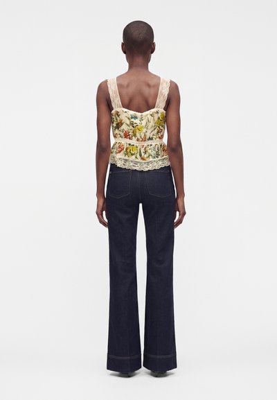 Femme debout, dos à la caméra, portant un top sans manches en dentelle fleurie et un jean large sombre sur un fond uni.