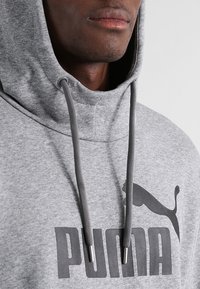 Šedá mikina s kapucí a stahovací šňůrkou, s velkým černým potiskem "PUMA" a logem. Měkká tkanina s hladkou texturou.