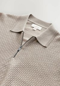 Suéter de punto beige con un patrón texturizado estructurado, cuello corto y una cremallera de un cuarto de longitud con un tirador negro.
