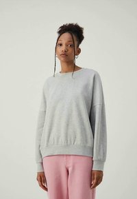 Jovem mulher de cabelo encaracolado, vestindo um sweatshirt cinza de mangas longas e calças rosas, de pé diante de um fundo claro e simples.