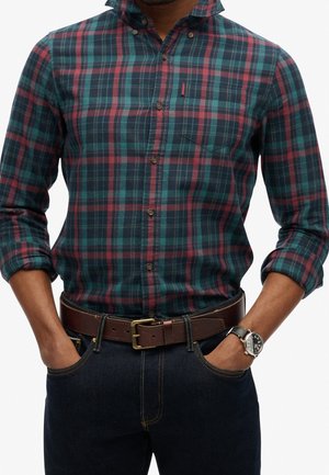 Mężczyzna noszący turkusowo-granatowo-czerwoną koszulę w kratę z podwiniętymi rękawami, ciemne jeansy, brązowy pasek i czarny zegarek na skórzanym pasku.