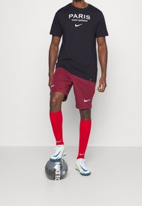 Czarna koszulka z napisem "PARIS SAINT-GERMAIN", bordowe spodenki, czerwone skarpety i białe/kobaltowe korki stojące na czarnej piłce nożnej.