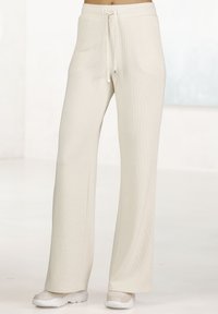 Helle beigen, gerippten Lounge-Hosen mit einem Kordelzugbund. Lockerer Schnitt mit geraden Beinen, kombiniert mit weißen Sportschuhen auf einem neutralen Hintergrund.