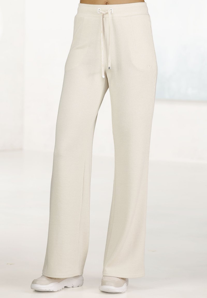 Helle beigen, gerippten Lounge-Hosen mit einem Kordelzugbund. Lockerer Schnitt mit geraden Beinen, kombiniert mit weißen Sportschuhen auf einem neutralen Hintergrund.