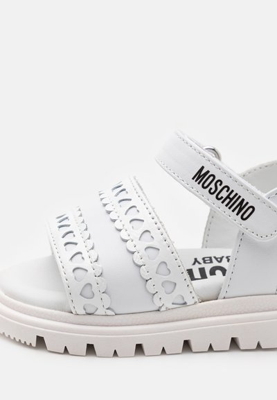 White sandals with heart-shaped cutouts, scalloped accents, and a thick white sole. Adjustable strap features "MOSCHINO" branding.