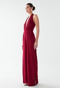 Mono burdeos con cuello halter y escote en V profundo, cintura ajustada y corte de pierna ancha. Presenta aberturas laterales y una textura con un brillo sutil.