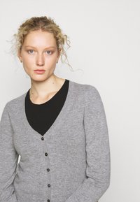 Grå cardigan laget av myk strikket stoff, med V-hals, lange ermer og brune knapper foran. Svart skjorte under.