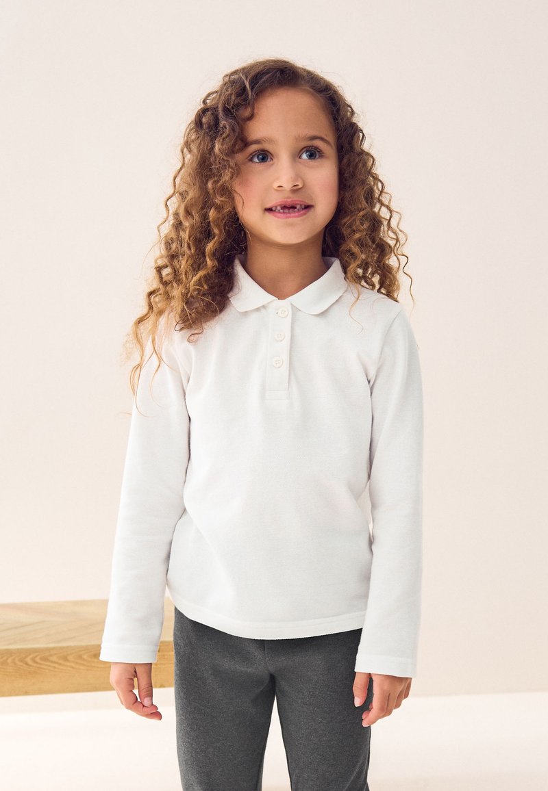 Junges Mädchen mit lockigem Haar, das ein weißes Langarm-Poloshirt und graue Hose trägt, steht drinnen vor einem einfarbigen hellen Hintergrund.