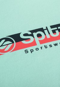 Helles mintgrünes Gewebe mit einem großen Grafikdesign in Schwarz und Rot. Der Text lautet prominent "Spit" und "Sportswear".