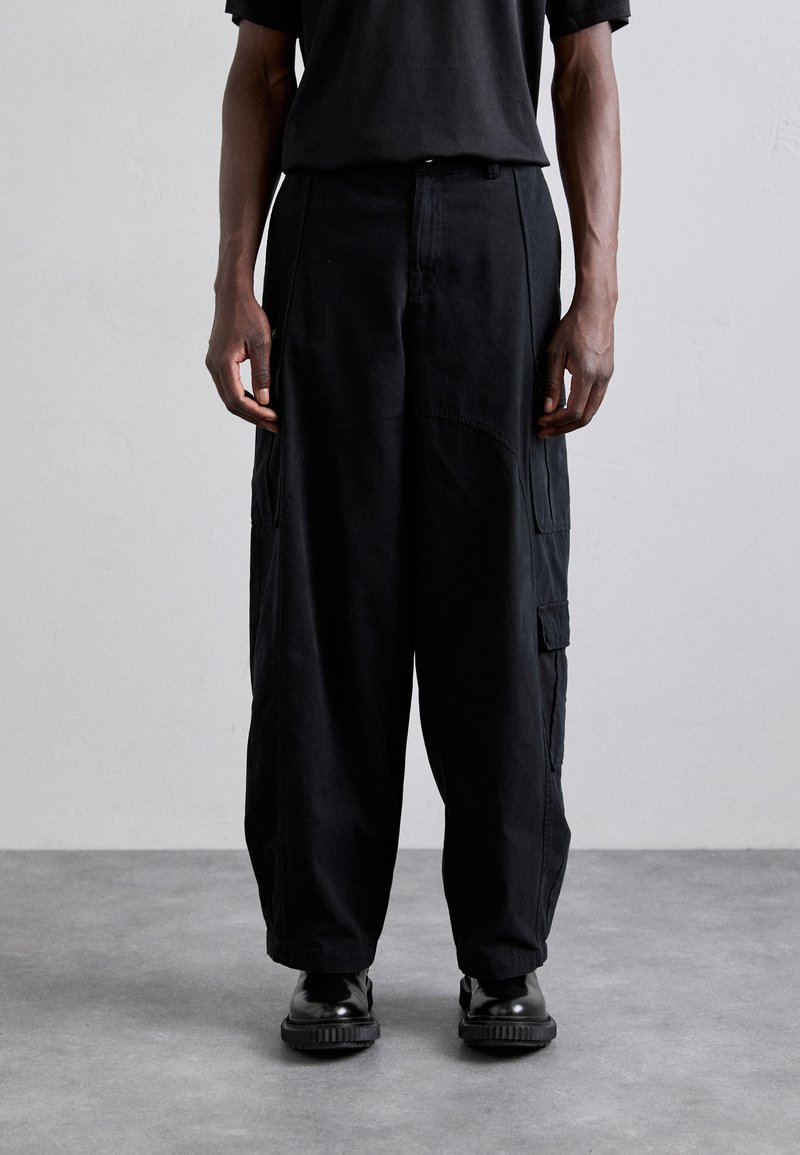 Svarte cargo-bukser med løs passform, justerbart liv, sidelommer og en glatt tekstur, kombinert med svarte sko.