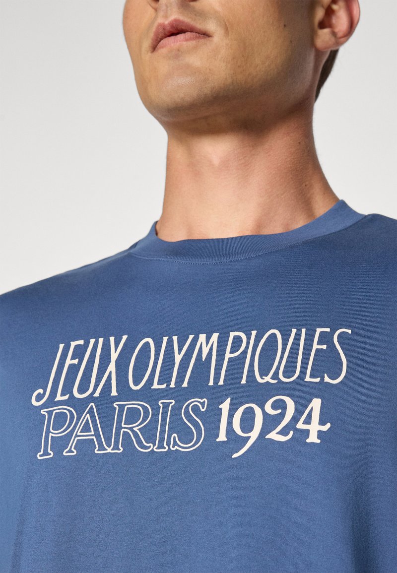 T-shirt en coton bleu avec l'inscription "JEUX OLYMPIQUES PARIS 1924" en texte blanc. Design classique à col rond, texture lisse et coupe décontractée.