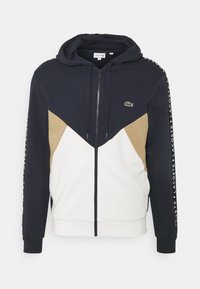Námořnický modrý, béžový a bílý zip-up mikina s kapucí, předními kapsami a logem Lacoste na hrudi a tištěným textem podél rukávů.