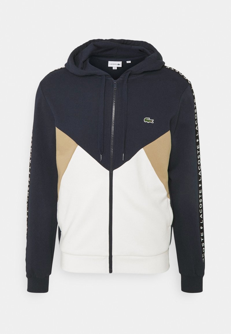 Námořnický modrý, béžový a bílý zip-up mikina s kapucí, předními kapsami a logem Lacoste na hrudi a tištěným textem podél rukávů.