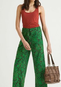 Top rosso senza maniche abbinato a pantaloni larghi verdi con motivo floreale. La modella tiene una borsa marrone testurizzata con dettagli.