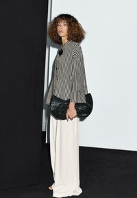 Sac en bandoulière noir tissé, haut rayé à manches longues noir et blanc, large pantalon crème, se tenant devant un fond minimaliste.