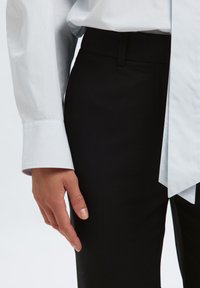 Camisa blanca de manga larga con cuello puntiagudo, combinada con pantalones negros ajustados. La tela parece suave y tiene un aspecto limpio y bien ajustado.