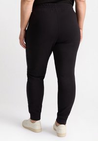 Schwarze Stretch-Jogginghosen mit elastischem Bund, schlankem Schnitt und gerippten Säumen, kombiniert mit weißen Sportschuhen.