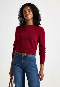 Rød cashmere sweater med lange ærmer; parret med højtaljede blå denimjeans og et brunt læderbælte. Simpel, klassisk design.