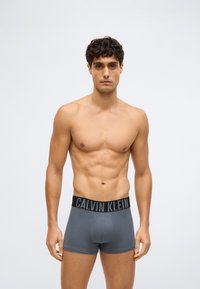 Kails vīrietis ar izcirtu tumšu matu stāv pelēkā gaismas fona priekšā, mugurā ģērbtām pelēkām Calvin Klein bokseru biksēm.