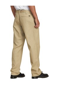 Pantalones de trabajo de algodón en beige, con corte recto, bolsillos traseros y una pequeña etiqueta con logo en la cintura. Llevados con calzado negro.