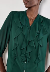 Blusa in chiffon verde con ampie ruches sul davanti, chiusura con bottoni e maniche a tre quarti. Texture liscia e morbido drappeggio.