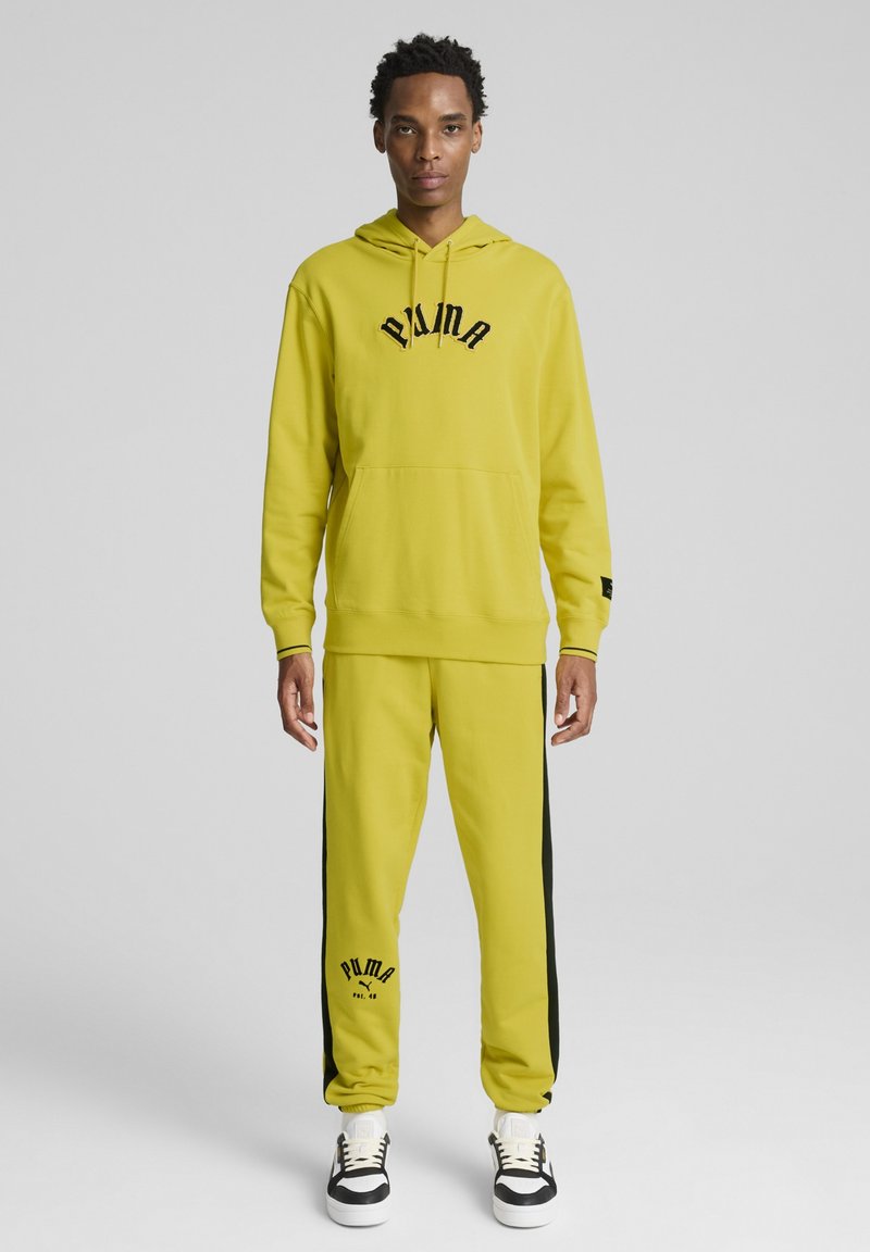 Gul huvtröja med svart text, kombinerad med matchande gula joggers med svarta sidostreck och svart logotyp. Komposit design för sportkläder.