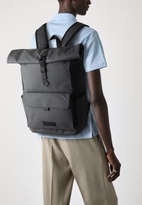 Black roll-top backpack with adjustable straps, front flap pocket, and smooth texture. Worn over a light blue shirt and beige trousers.