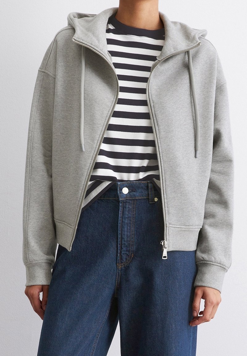 Personne portant un sweat à capuche gris zippé par-dessus un t-shirt rayé noir et blanc, rentré dans un jean bleu foncé taille haute.