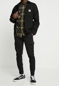 Man som bär en svart jacka, grön och svart rutad skjorta, svarta cargobyxor och svart-vita sneakers, stående med en hand i fickan.