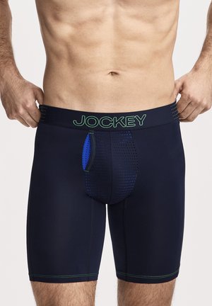 Männlicher Oberkörper trägt dunkelblaue Jockey Boxershorts mit perforiertem Frontbeutel, hält den Bund auf beiden Seiten vor einfarbigem Hintergrund.
